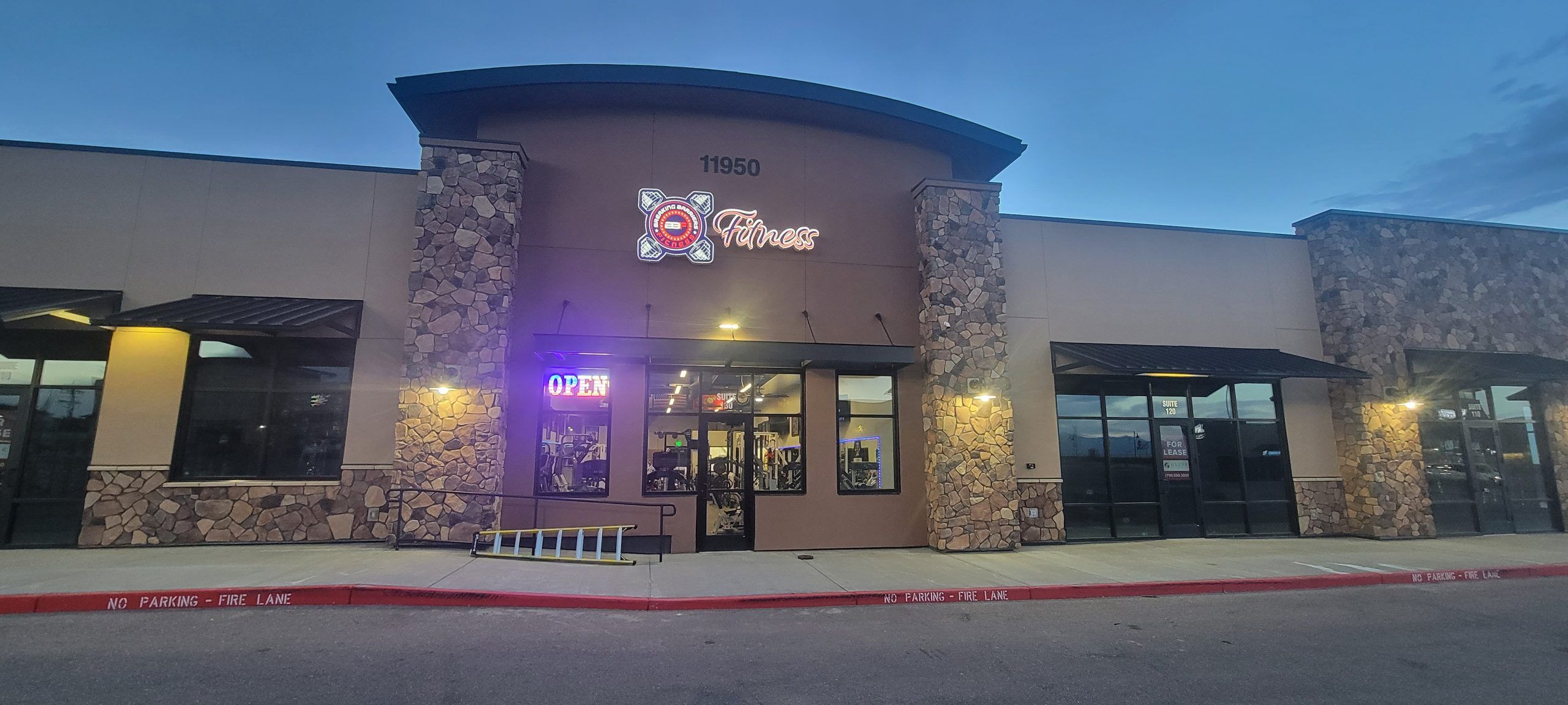 Breaking Barriers Fitness storefront at dusk, with the 11950 address sign, lit logo and OPEN sign on Tourmaline Drive in Peyton, Colorado
